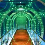 A narrow, wooden hallway with a glass railing. Surrounded by green lights.