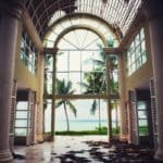 Inside a building with tall open ceiling. Looking out is a giant glass wall that looks out to a body of water with palm trees.