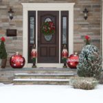 A brown front door with a christmas wreath hanging on it. On the front porch is two toy solder and giant red ornaments on both side.