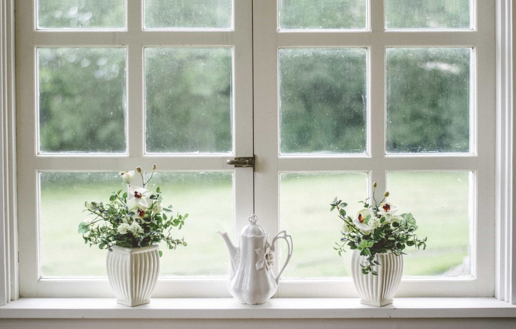 A white window frame holds two pots with greenery inside and between them is a white teapot.