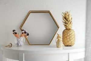 Golden framed hexagon mirror sits on a white table against a white wall. On the table is a glass is makeup brushes, an eyelash curler, and two golden pineapples one large and one small.