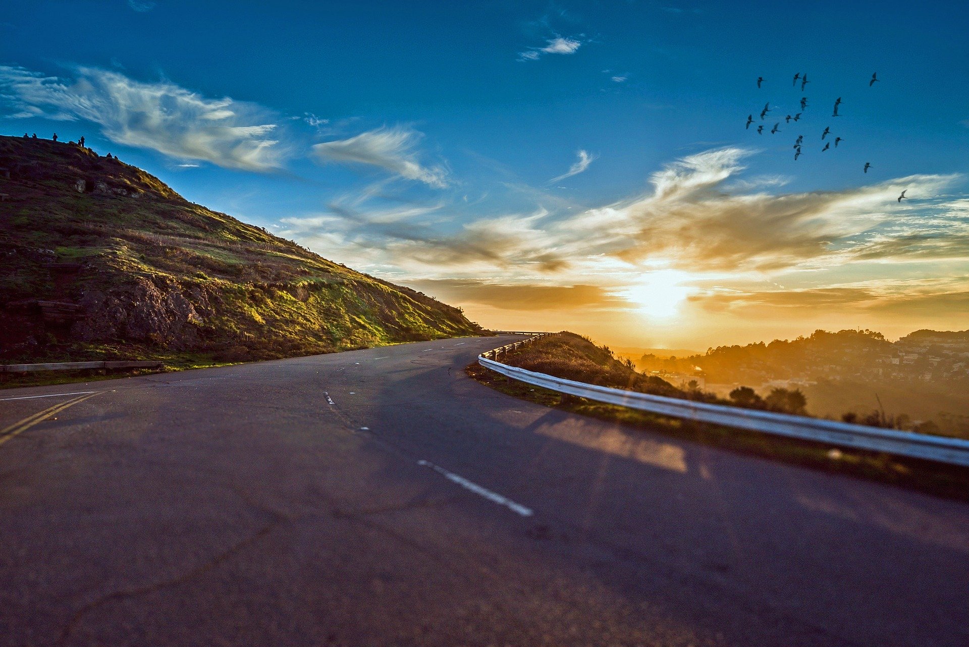 A bright blue sky with an orange sun, is slightly covered by thin clouds. The sun illuminates a dark road.
