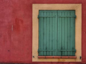 Green hurricane shutters cover a window of a red building wall.