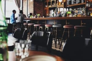 An overview of a bar. At the wooden bar is a line of cloth covered stools. Two people sit at the bar.