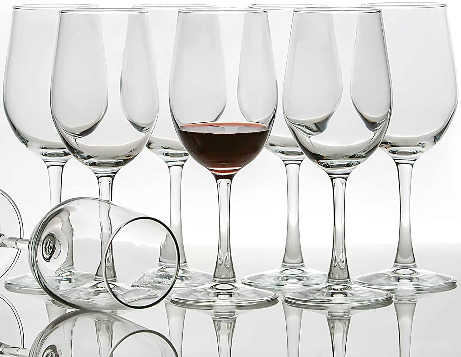 8 All-purpose wine glasses sit on a white counter with a white background. The glass in the front middle is filled with red wine. Another glass is fallen over to see inside the top of the glass.