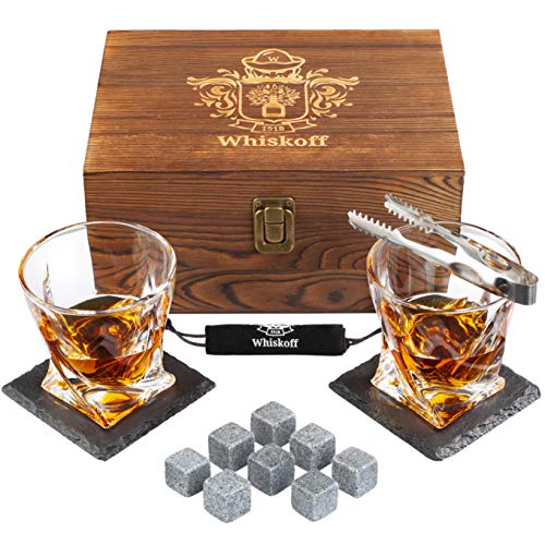 A brown wooden box with a golden latch and golden wording that reads "Whiskoff" sits in the back of the image. Two half full crystal whiskey glasses sit on two dark square, gray stone coasters. One of the glasses has a small silver tong sitting on top of the glass rim. In front of the two glasses are 8 cubed small gray stones.