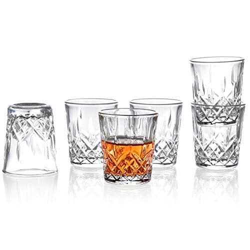 6 crystal whiskey shot glasses sit against a white background. One glass in the front is half full of whiskey.